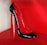 A high-heeled shoe with a glossy black exterior and red interior, featuring a unique silver stiletto heel. The shoe is displayed against a bright red background, highlighting its elegant and sophisticated design.