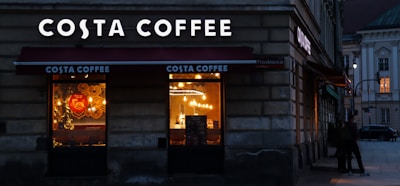 A Costa Coffee shop is situated on a corner of a street with illuminated signage and warm light emanating from the windows. The shop's windows display decorative elements and are partially obscured by reflections. Outside, the street is dimly lit, creating a cozy atmosphere.