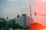 A multi-story building with a large telecommunications tower on top is seen amidst a busy urban setting. Traffic lights and a few motor vehicles are present, alongside some people wearing helmets. There are trees in the background and a prominent red light leak effect partially obscuring the image.