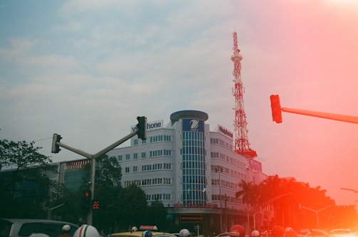 A multi-story building with a large telecommunications tower on top is seen amidst a busy urban setting. Traffic lights and a few motor vehicles are present, alongside some people wearing helmets. There are trees in the background and a prominent red light leak effect partially obscuring the image.