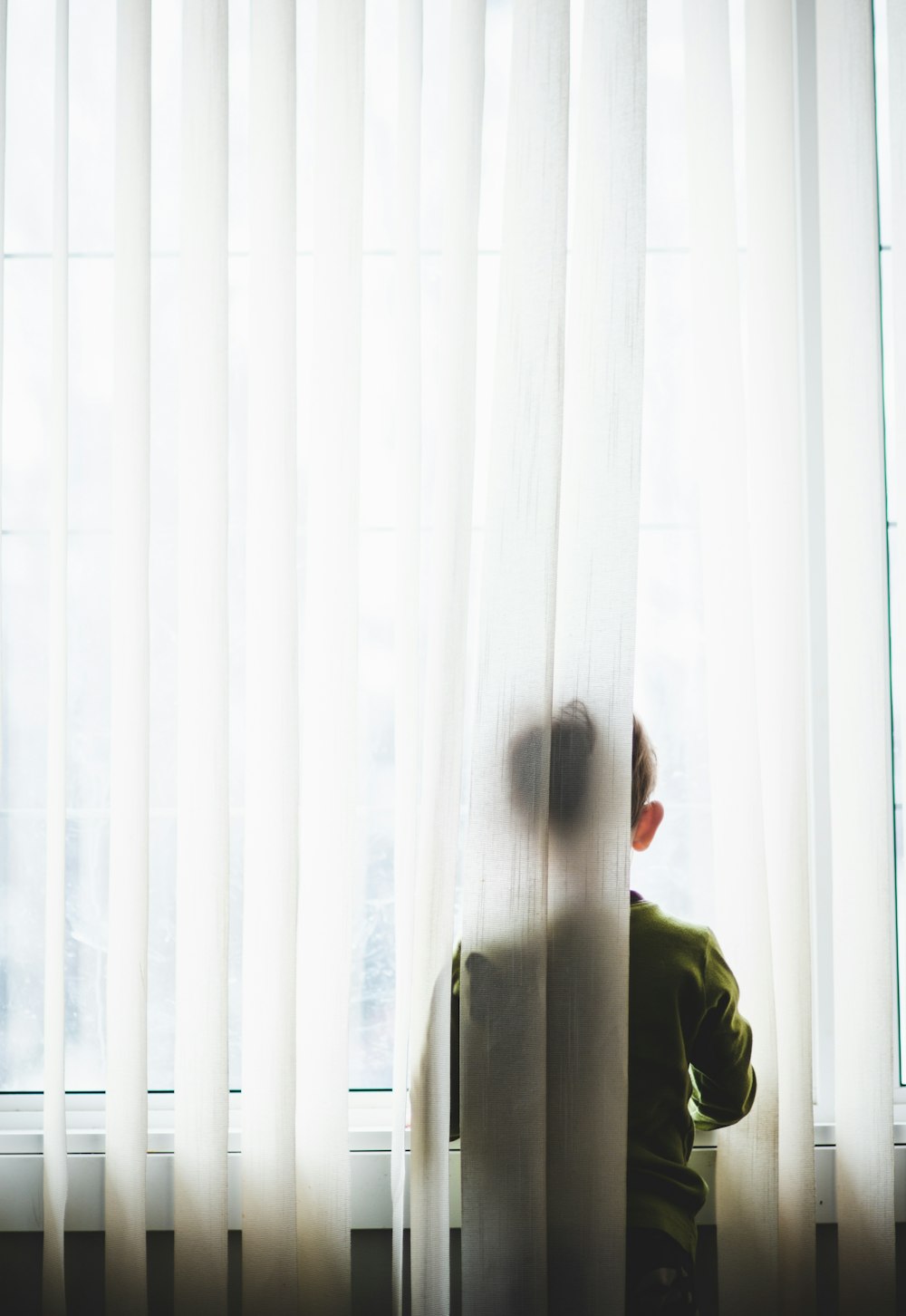 Hombre en abrigo verde de pie cerca de la cortina blanca de la ventana