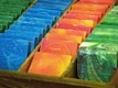 Several rectangular bars of soap are neatly arranged in rows. The soaps are in a variety of vibrant colors including blue, red, yellow, orange, and green. Each bar appears to have embossed designs or writing on them, suggesting they are decorative or artisanal.