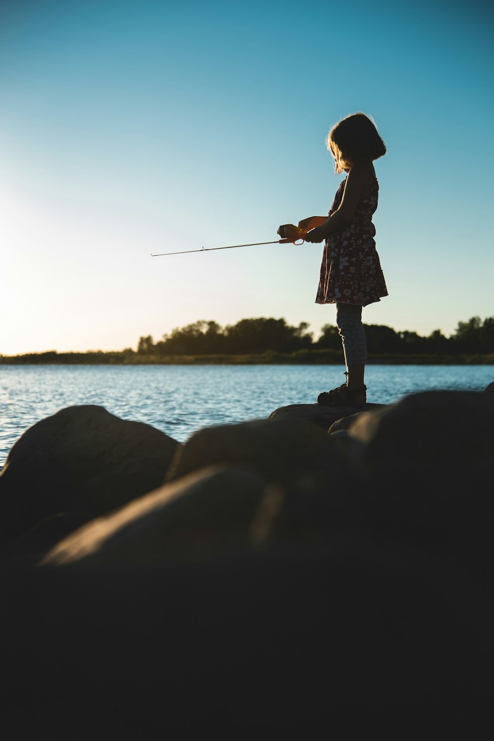 Femme en robe blanche et noire tenant une canne à pêche debout sur un rocher près d’un plan d’eau