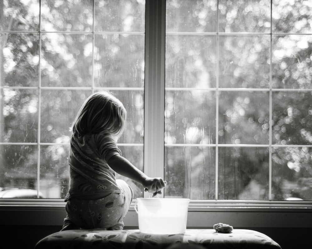 grayscale photo of woman sitting on table looking at the window