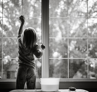 grayscale photo of girl in stripe shirt and pants standing by the window