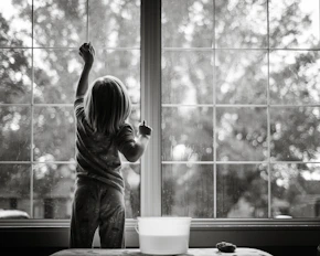 grayscale photo of girl in stripe shirt and pants standing by the window