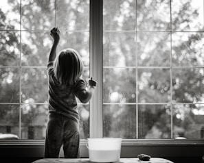 grayscale photo of girl in stripe shirt and pants standing by the window