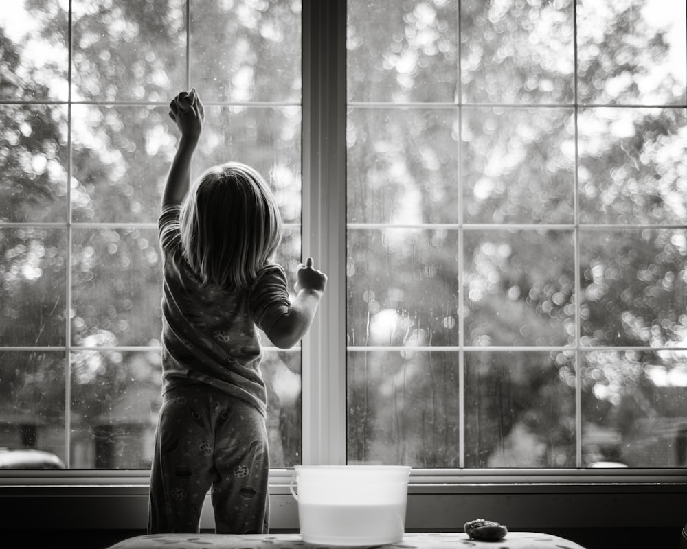 Foto en escala de grises de una chica con camisa a rayas y pantalones de pie junto a la ventana