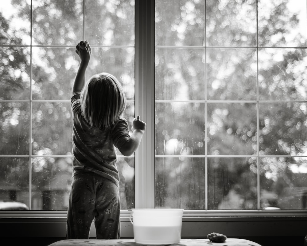 Graustufenfoto eines Mädchens in gestreiftem Hemd und Hose, das am Fenster steht