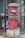 A red cylindrical post box stands in front of a black iron gate. The post box has a domed top and is mounted on a concrete base. There is text on the post box indicating 'Cumballa Hill 400026'. A car tire and part of a corrugated metal sheet are visible in the background.