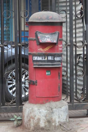 A red cylindrical post box stands in front of a black iron gate. The post box has a domed top and is mounted on a concrete base. There is text on the post box indicating 'Cumballa Hill 400026'. A car tire and part of a corrugated metal sheet are visible in the background.