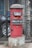 A red cylindrical post box stands in front of a black iron gate. The post box has a domed top and is mounted on a concrete base. There is text on the post box indicating 'Cumballa Hill 400026'. A car tire and part of a corrugated metal sheet are visible in the background.