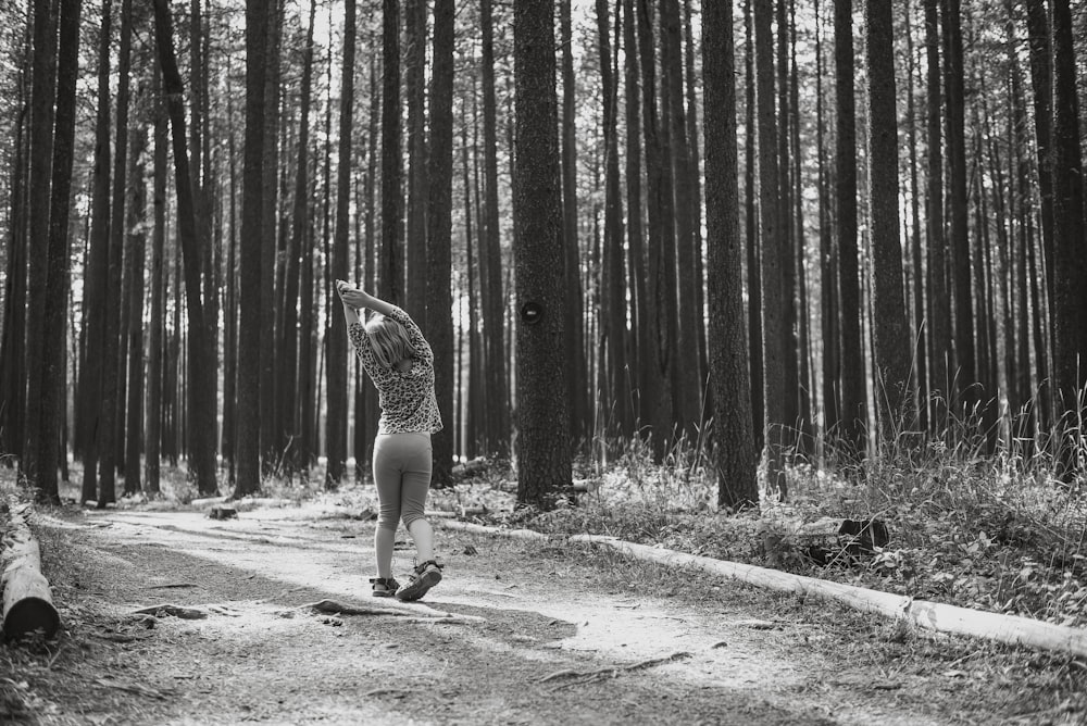 grayscale photo of woman in dress walking on pathway in between trees