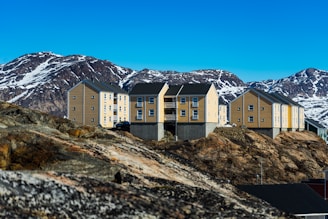 A group of modern, light-brown buildings with dark roofs situated on rocky terrain. Behind the structures, a backdrop of snow-capped, rugged mountains rises under a clear blue sky. The architecture is minimalist, blending with the natural landscape.