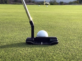 A close-up shot of a golf putter poised next to a golf ball on a well-manicured green. The focus is on the putter and the ball, with the flag in the background amidst a few other golf balls.