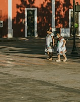 Two women walk together on a sunlit street in front of a vibrant red building. Both are wearing traditional, colorful embroidered dresses. Their expressions suggest companionship and support.