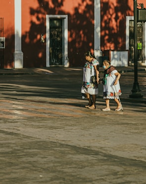 Two women walk together on a sunlit street in front of a vibrant red building. Both are wearing traditional, colorful embroidered dresses. Their expressions suggest companionship and support.
