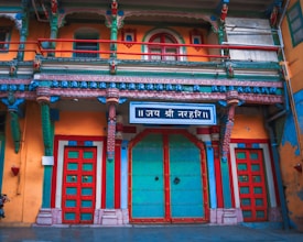 The image displays a brightly colored, intricately detailed facade of a building with ornate designs. The vibrant colors include turquoise, red, orange, and green, forming a visually striking composition. Multiple doors and windows feature colorful frames and patterns, with a prominent sign written in a script above the central door.