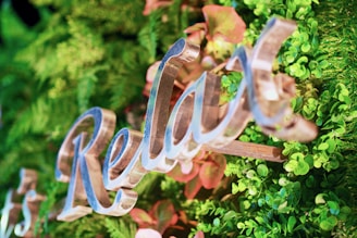 The image features a close-up view of decorative, cursive lettering spelling 'Relax' against a vibrant backdrop of lush green foliage with some reddish hues from leaves. The letters have a shiny, metallic appearance and are artistically integrated into the natural setting.
