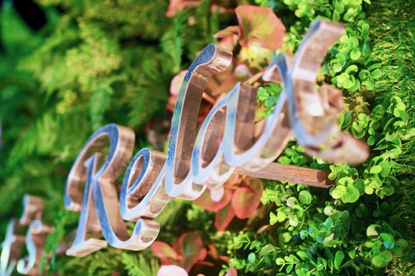 The image features a close-up view of decorative, cursive lettering spelling 'Relax' against a vibrant backdrop of lush green foliage with some reddish hues from leaves. The letters have a shiny, metallic appearance and are artistically integrated into the natural setting.