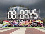 A large countdown display features '00 DAYS' made from giant white numeric panels supported by a metal framework. Below, a vibrant mural depicts colorful cartoon characters, musical notes, and whimsical designs. Logos and text promoting the Eurovision Song Contest in Rotterdam 2021 appear, highlighted by bold colors against a backdrop of dark, stormy clouds.