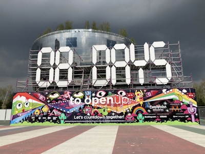 A large countdown display features '00 DAYS' made from giant white numeric panels supported by a metal framework. Below, a vibrant mural depicts colorful cartoon characters, musical notes, and whimsical designs. Logos and text promoting the Eurovision Song Contest in Rotterdam 2021 appear, highlighted by bold colors against a backdrop of dark, stormy clouds.