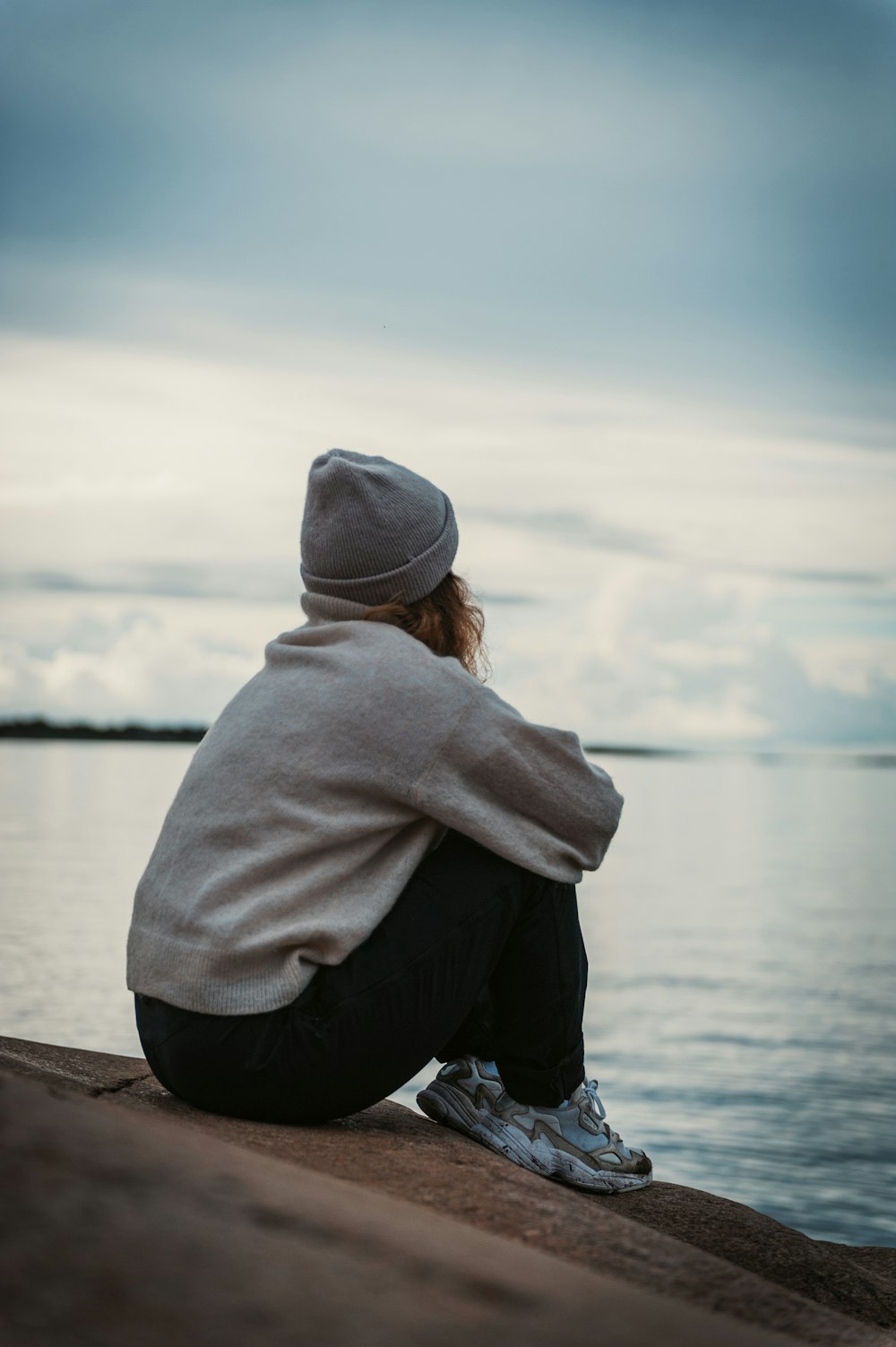 Persona con sudadera con capucha gris y pantalones negros sentada en un muelle de madera marrón durante el día