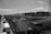 A large industrial area with multiple storage tanks and railway tracks leading into the facility. Several trucks are parked along a road on the left side. There are numerous pipes and infrastructure supporting the tanks, as well as communication towers and small structures scattered throughout. The sky is partly cloudy and the image is in grayscale.