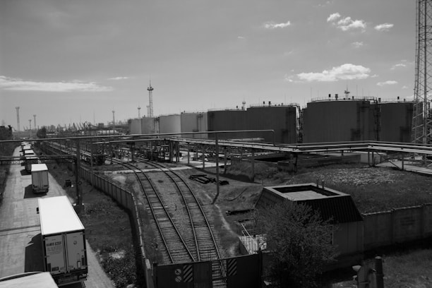 A large industrial area with multiple storage tanks and railway tracks leading into the facility. Several trucks are parked along a road on the left side. There are numerous pipes and infrastructure supporting the tanks, as well as communication towers and small structures scattered throughout. The sky is partly cloudy and the image is in grayscale.