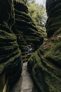 A narrow stone pathway winds between towering moss-covered rock formations. A person wearing a gray hoodie stands on the path, looking through binoculars. Dense vegetation and trees are visible above, contributing to an overall sense of lushness and seclusion.