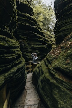 A narrow stone pathway winds between towering moss-covered rock formations. A person wearing a gray hoodie stands on the path, looking through binoculars. Dense vegetation and trees are visible above, contributing to an overall sense of lushness and seclusion.