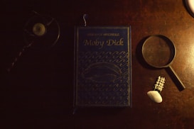 A hardcover book titled Moby Dick by Herman Melville is placed on a wooden table. The book has a dark blue cover with an illustration of a whale and waves. To the left, there is a candle holder with a small candle, and to the right, there is a magnifying glass, a piece of white coral, and a shell.