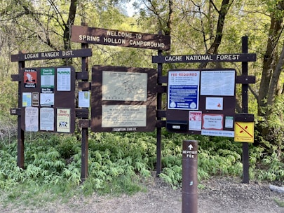 A large informational signboard with multiple notices and maps, located at a campground entrance. Surrounded by dense green foliage, the signs provide details about camping regulations, fees, and safety tips in the area.