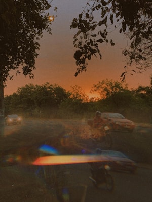 The image captures a twilight scene with the sun setting in the background, casting an orange glow across the sky. Trees and foliage are silhouetted against the vibrant horizon. In the foreground, a road can be seen with a car and a person on a motorcycle moving forward. The image is overlaid with a prism effect, creating a colorful and dreamy atmosphere.