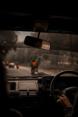 View from the inside of a car driving on a road. The driver&rsquo;s hand is on the steering wheel. Several motorcyclists with orange vests are visible ahead on the road. The image is somewhat dark with a moody atmosphere, likely during dawn or dusk. The rearview mirror reflects a portion of the driver's face.
