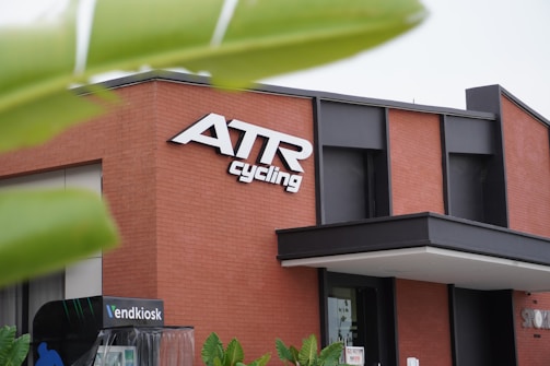 A modern building with a red brick facade and large black-framed windows. Prominent signage on the building displays 'ATR Cycling' in bold white letters. There are green leaves partially obscuring the view in the foreground, adding a natural contrast to the architectural scene. Additional smaller signs or structures are visible near the base of the building.