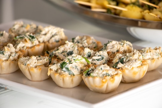 A platter of small pastry cups filled with a creamy, textured topping, likely containing spinach and herbs. They are neatly arranged in rows on a rectangular white plate. In the background, a blurred section of another dish is visible.
