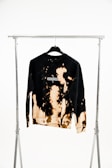 A black sweatshirt with a unique bleach tie-dye pattern hanging on a black hanger attached to a metal garment rack. The design features light brown and beige splashes across the fabric, with the word 'enigma' printed on the chest.