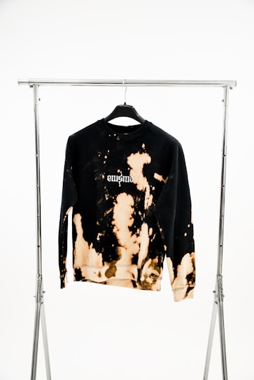 A black sweatshirt with a unique bleach tie-dye pattern hanging on a black hanger attached to a metal garment rack. The design features light brown and beige splashes across the fabric, with the word 'enigma' printed on the chest.