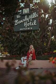 A woman with long blonde hair is sitting on a low wall in front of lush greenery. She is wearing a long floral skirt, white shoes, and a red top, holding a green purse. Above her is a sign for the Beverly Hills Hotel, surrounded by large tropical plants.
