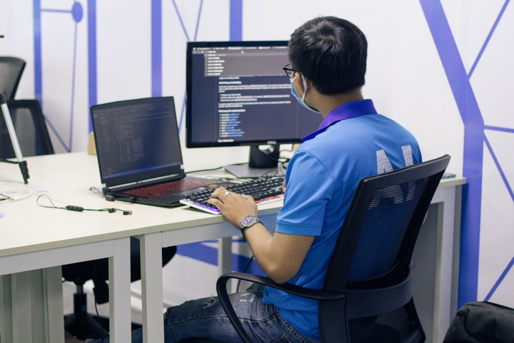 Ragazzo in maglietta blu che si siede sulla sedia nera di rotolamento dell'ufficio davanti al computer