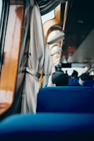 A view inside a vehicle, likely a bus, showcasing the back of blue seats with a few passengers visible, some wearing face masks. Wooden paneling and light-colored curtains adorn the windows, and a sense of calmness pervades the setting.