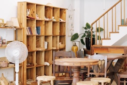 A cozy wooden interior featuring a large wooden shelf with open compartments filled with books and small decorative items. A standing fan is positioned next to the shelf. The room includes a round wooden table surrounded by various wooden chairs. There is a white staircase with wooden railings on the right side, and potted plants add a touch of greenery to the space.