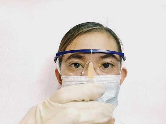 A person wearing protective gear including safety goggles, a surgical mask, and gloves is holding a syringe with a needle pointed upwards. The background is plain and light-colored, focusing attention on the person's face and the syringe.