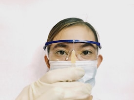 A person wearing protective gear including safety goggles, a surgical mask, and gloves is holding a syringe with a needle pointed upwards. The background is plain and light-colored, focusing attention on the person's face and the syringe.