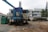 A construction site features large machinery with visible rust and signs of wear. The equipment includes a large blue machine with a tarp covering part of it and an adjacent green generator. In the background, there's a stack of concrete blocks, along with a white industrial building on the right. Some trees are seen to the right, providing a hint of greenery amidst the industrial setting.