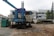 A construction site features large machinery with visible rust and signs of wear. The equipment includes a large blue machine with a tarp covering part of it and an adjacent green generator. In the background, there's a stack of concrete blocks, along with a white industrial building on the right. Some trees are seen to the right, providing a hint of greenery amidst the industrial setting.