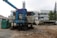 A construction site features large machinery with visible rust and signs of wear. The equipment includes a large blue machine with a tarp covering part of it and an adjacent green generator. In the background, there's a stack of concrete blocks, along with a white industrial building on the right. Some trees are seen to the right, providing a hint of greenery amidst the industrial setting.