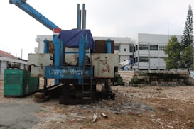 A construction site features large machinery with visible rust and signs of wear. The equipment includes a large blue machine with a tarp covering part of it and an adjacent green generator. In the background, there's a stack of concrete blocks, along with a white industrial building on the right. Some trees are seen to the right, providing a hint of greenery amidst the industrial setting.