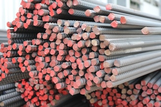 A large bundle of stacked metal rebars with rust-colored ends is shown. The rebars have a ribbed texture and are neatly arranged in parallel rows, extending to the background.